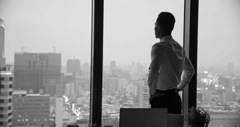 black and white photo of a man looking out of a large window of his office overlooking the rest of the town. Some buildings and high rises are visible in the background.