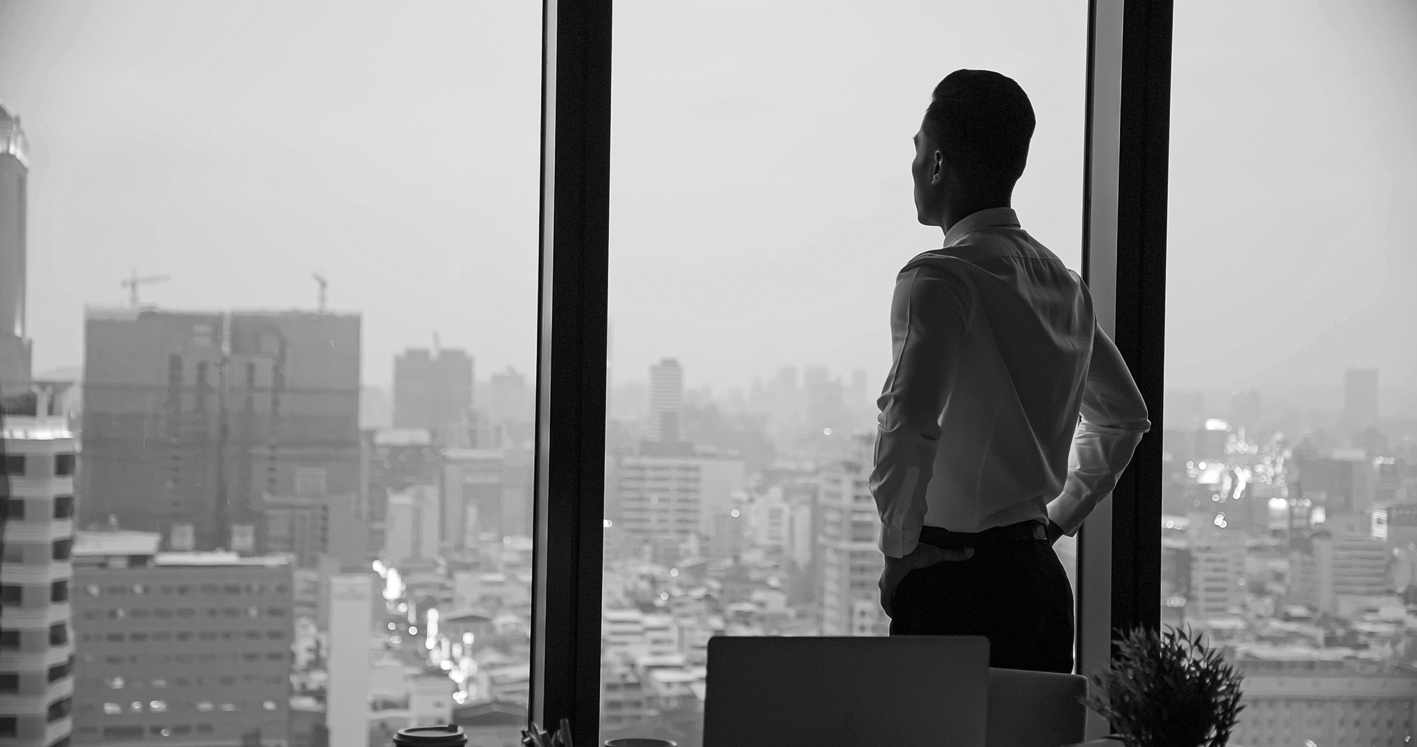black and white photo of a man looking out of a large window of his office overlooking the rest of the town. Some buildings and high rises are visible in the background.